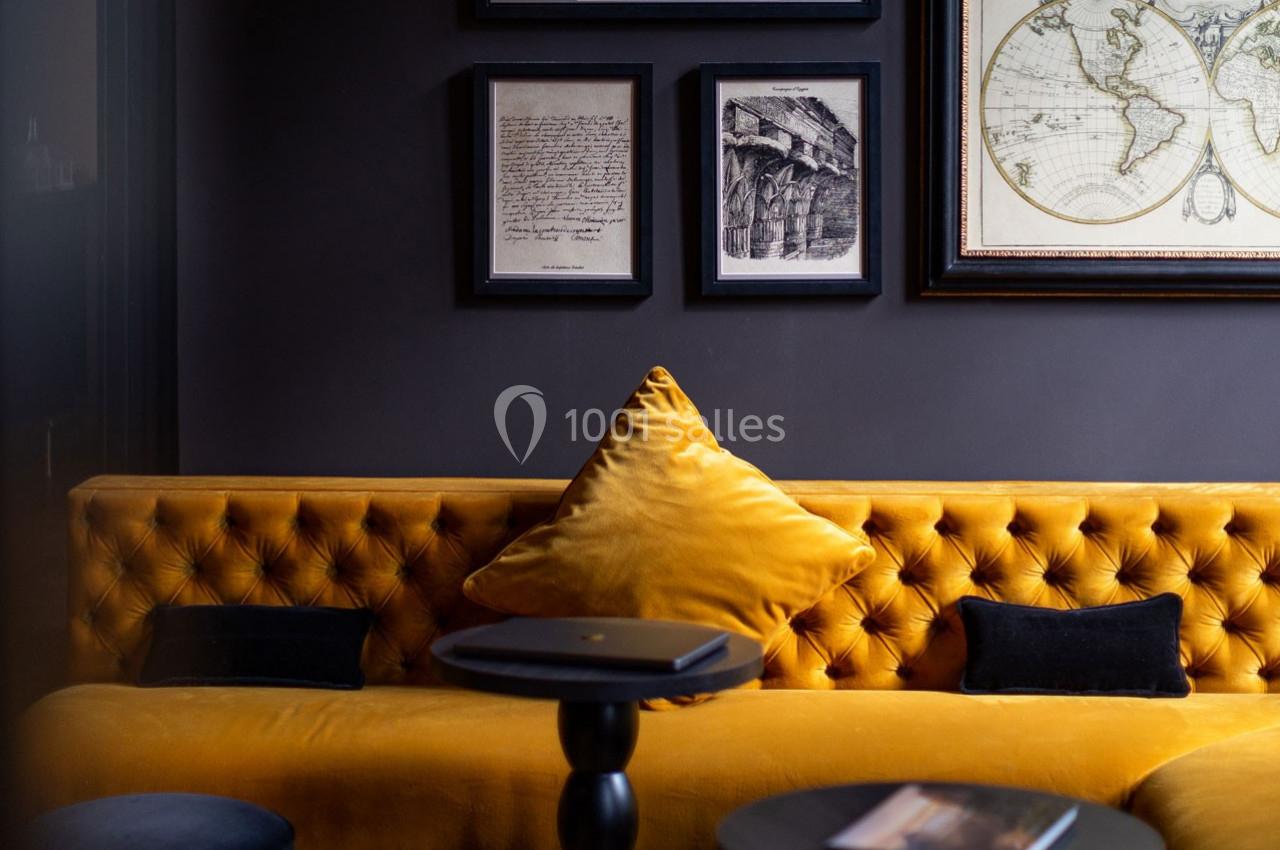 Canapé jaune moutarde capitonné avec coussins noirs, tables basses noires et cadres décoratifs accrochés au mur sombre.