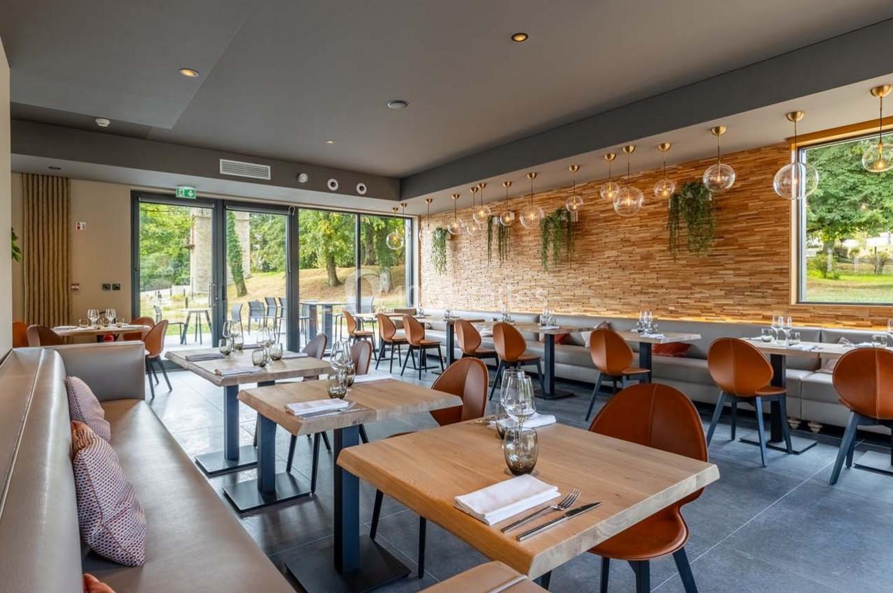 Salle de restaurant moderne avec tables en bois, chaises en cuir, grandes fenêtres et vue sur un espace vert.