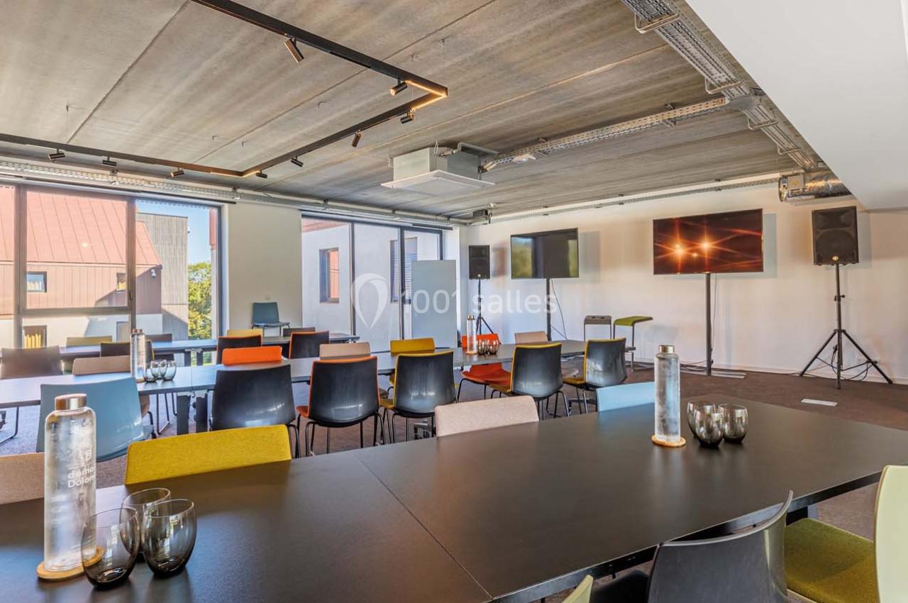 Salle de réunion moderne avec tables noires, chaises colorées, écrans, grandes fenêtres et éclairage au plafond.