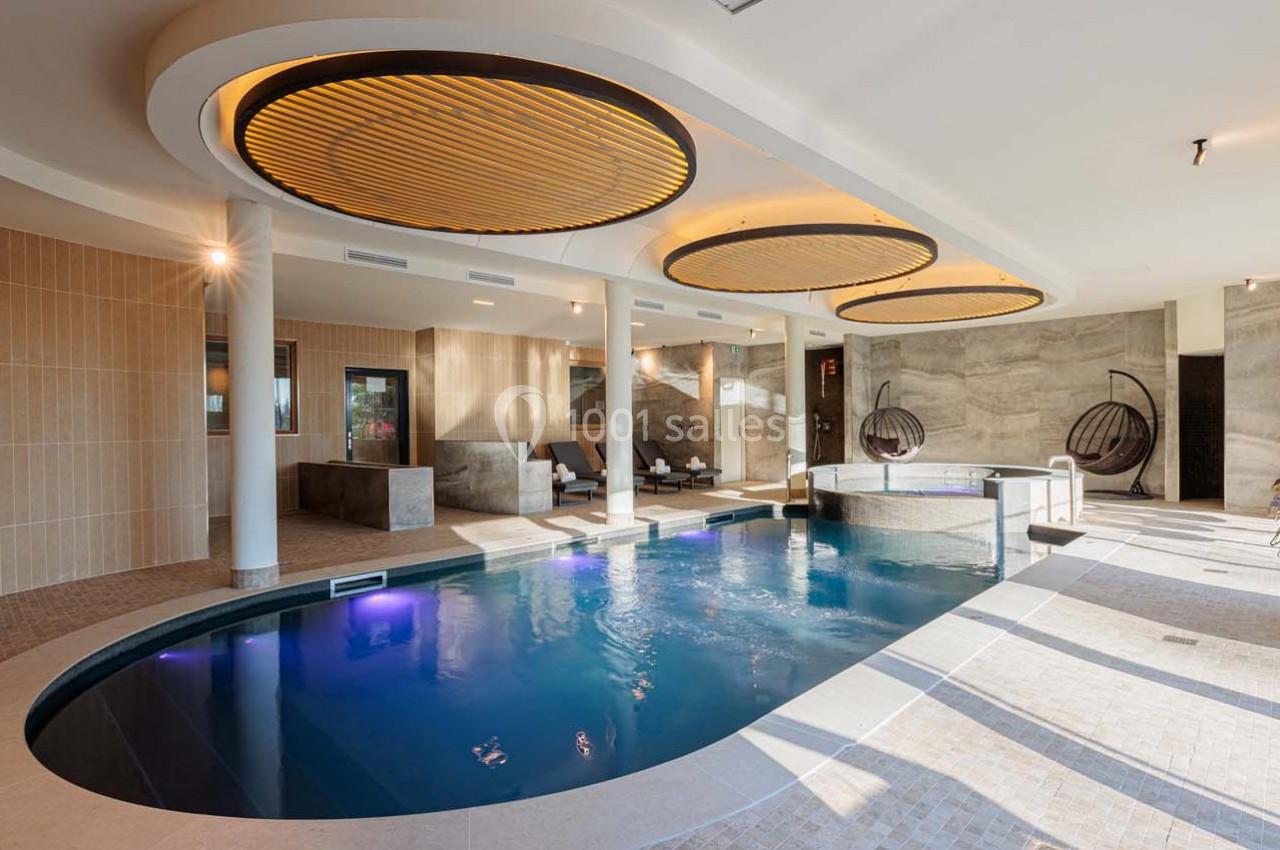 Piscine intérieure moderne avec éclairage tamisé, entourée de chaises longues et de murs en pierre beige.