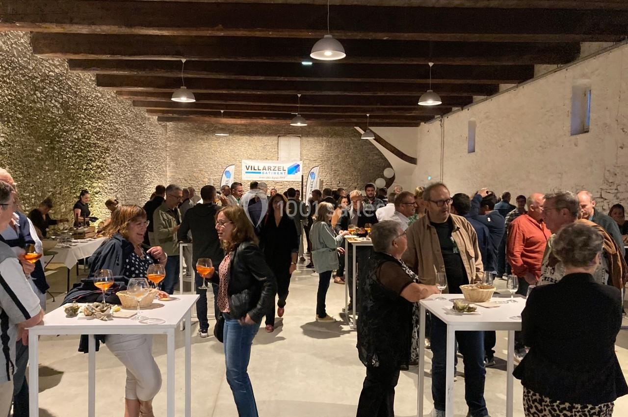 Des personnes discutent et partagent un apéritif dans une salle en pierre avec des tables hautes et un éclairage chaleureux.