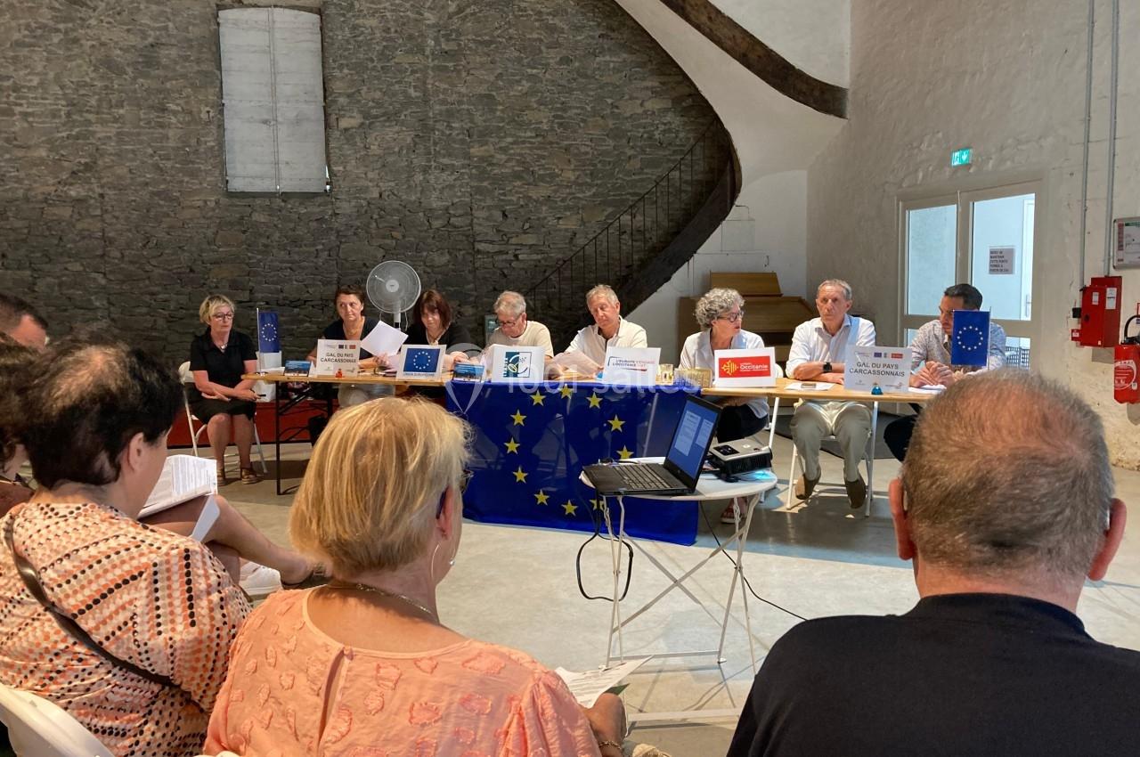 Un groupe de personnes assises à une table lors d'une réunion dans une salle avec un drapeau de l'Union européenne.