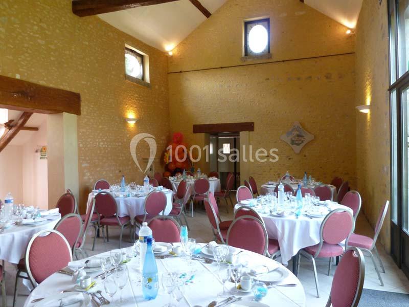 Salle de réception avec murs en pierre, tables rondes dressées et chaises rouges, éclairée par des fenêtres.