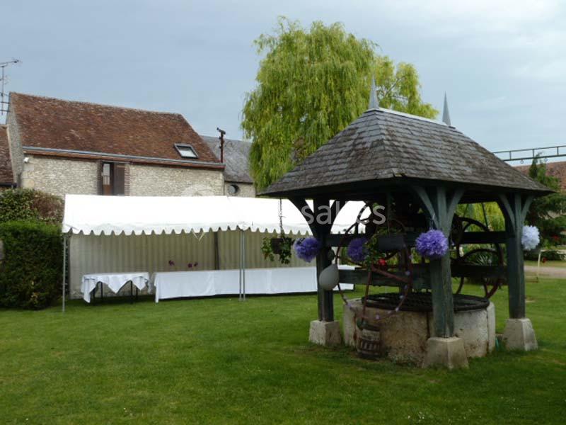 Puits ancien en pierre avec un toit en bois, devant une tente blanche installée sur une pelouse verte.