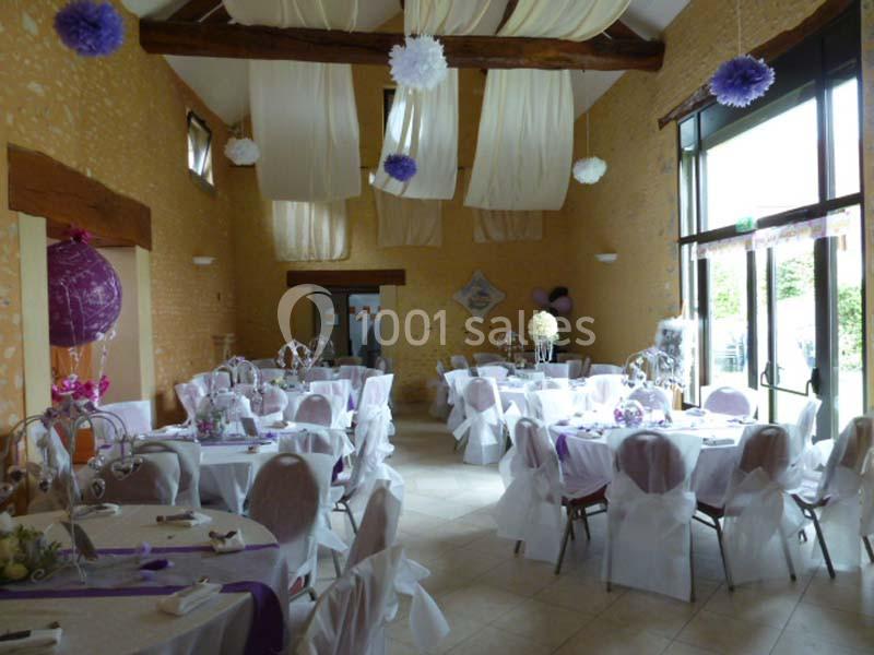 Salle de réception décorée avec des tables rondes, nappes blanches, chaises ornées et suspensions violettes et blanches.