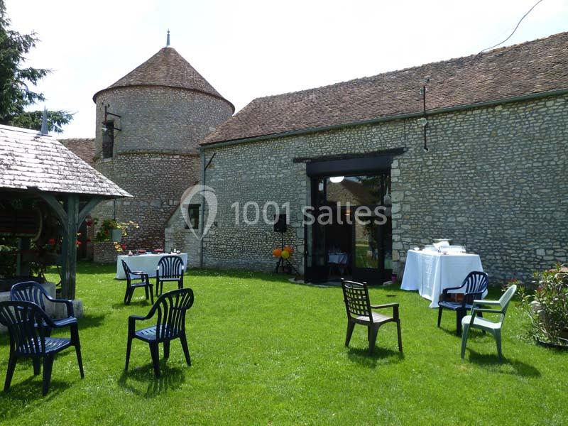 Cour verdoyante avec des chaises en plastique dispersées, une table dressée et un bâtiment en pierre en arrière-plan.