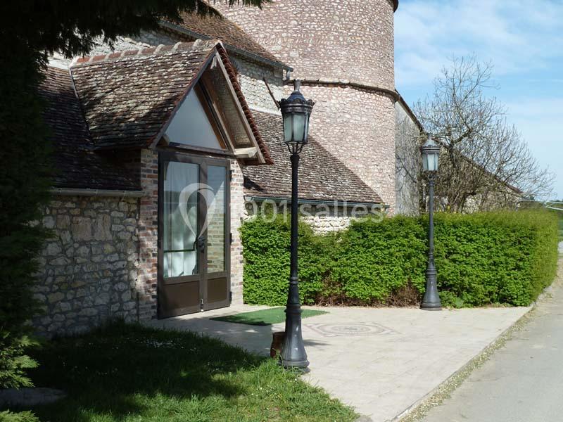 Entrée d'un bâtiment en pierre avec une porte vitrée, lampadaires noirs et haie verte sous un ciel dégagé.