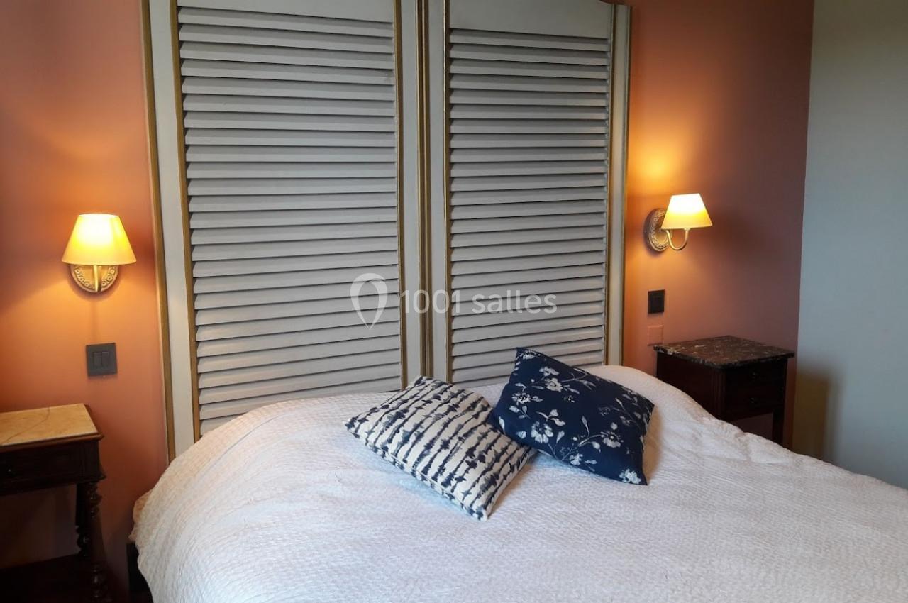 Lit double avec coussins décoratifs devant des portes en bois à persiennes, éclairé par deux appliques murales.