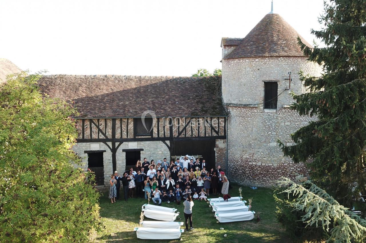Groupe de personnes rassemblées devant un bâtiment en pierre avec une tour et un toit en tuiles, entouré de verdure.