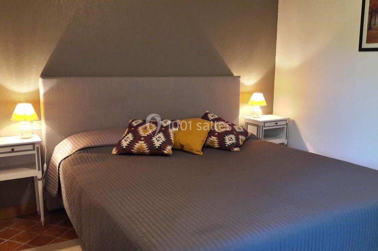 Lit double avec couvre-lit gris, coussins décoratifs colorés, deux lampes de chevet allumées et mur beige en arrière-plan.