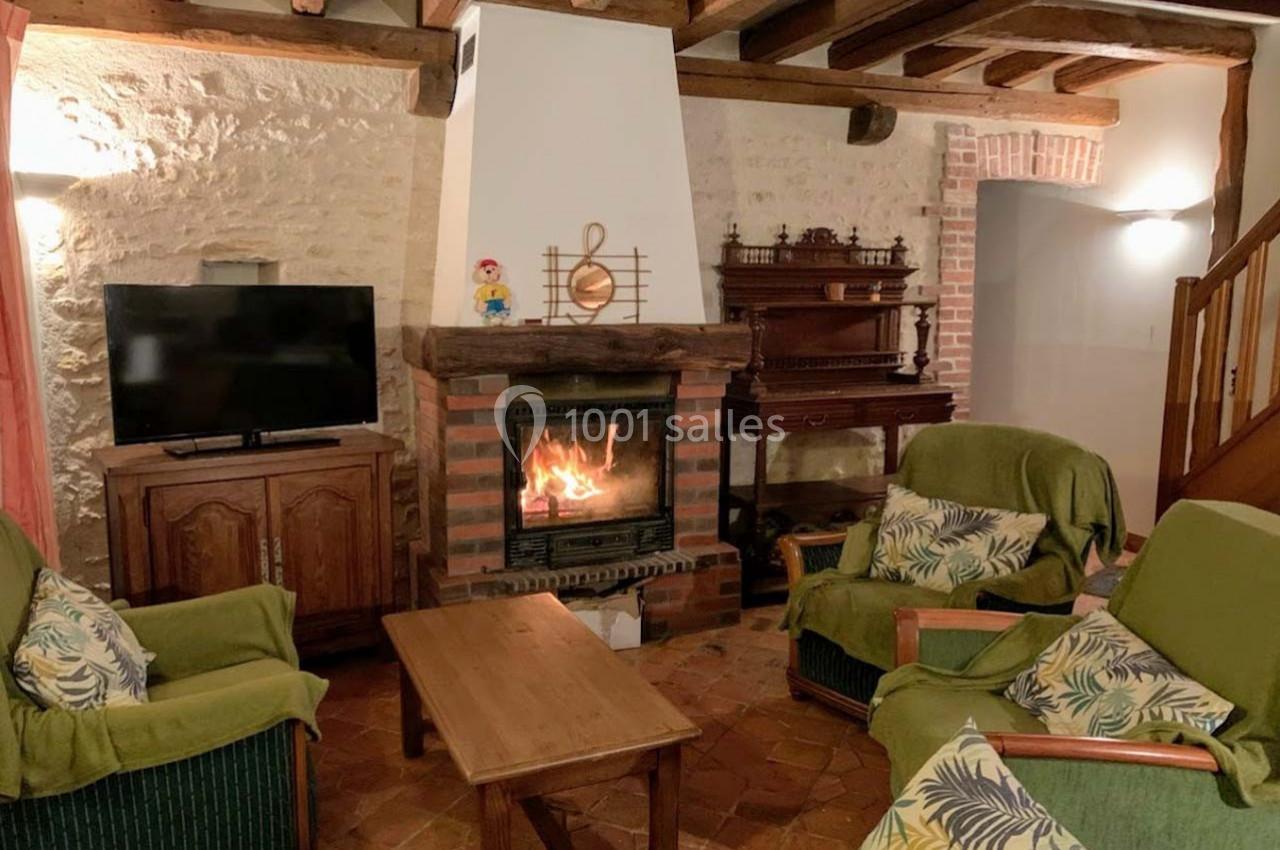 Salon chaleureux avec cheminée allumée, meubles en bois, fauteuils verts et murs en pierre apparente.