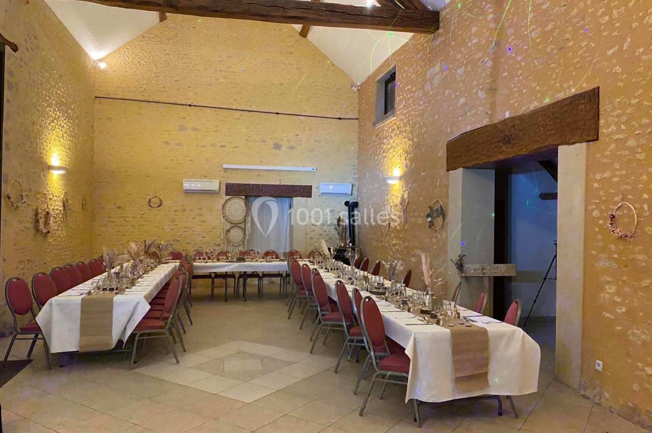 Salle de réception avec murs en pierre, poutres apparentes et tables dressées pour un repas, entourées de chaises rouges.