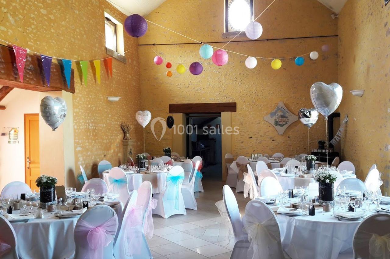 Salle décorée pour une réception avec tables dressées, chaises ornées de rubans colorés et guirlandes suspendues.