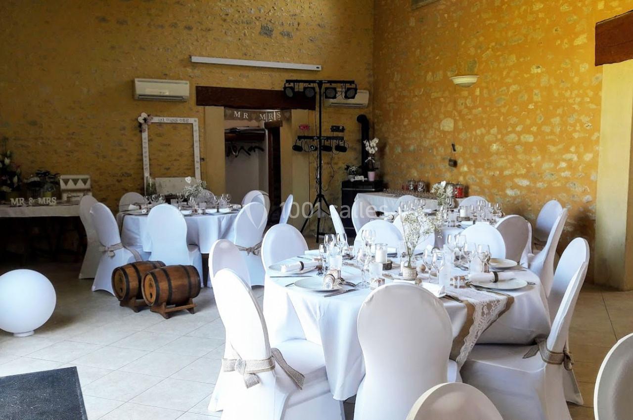 Salle de réception décorée pour un mariage, avec tables rondes dressées, chaises blanches et éléments rustiques.