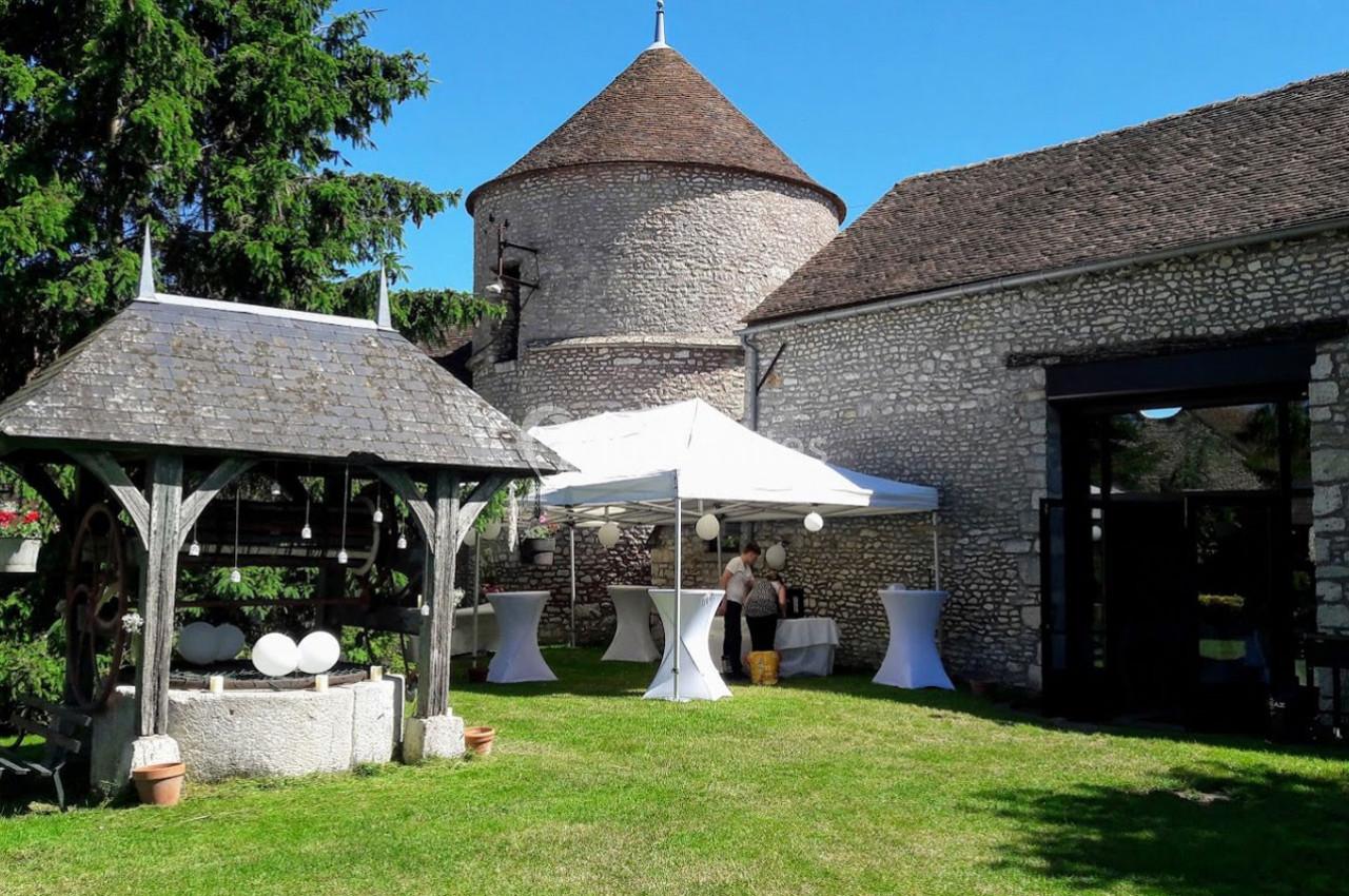 Cour extérieure avec pelouse, puits couvert, tente blanche et bâtiment en pierre sous un ciel bleu.