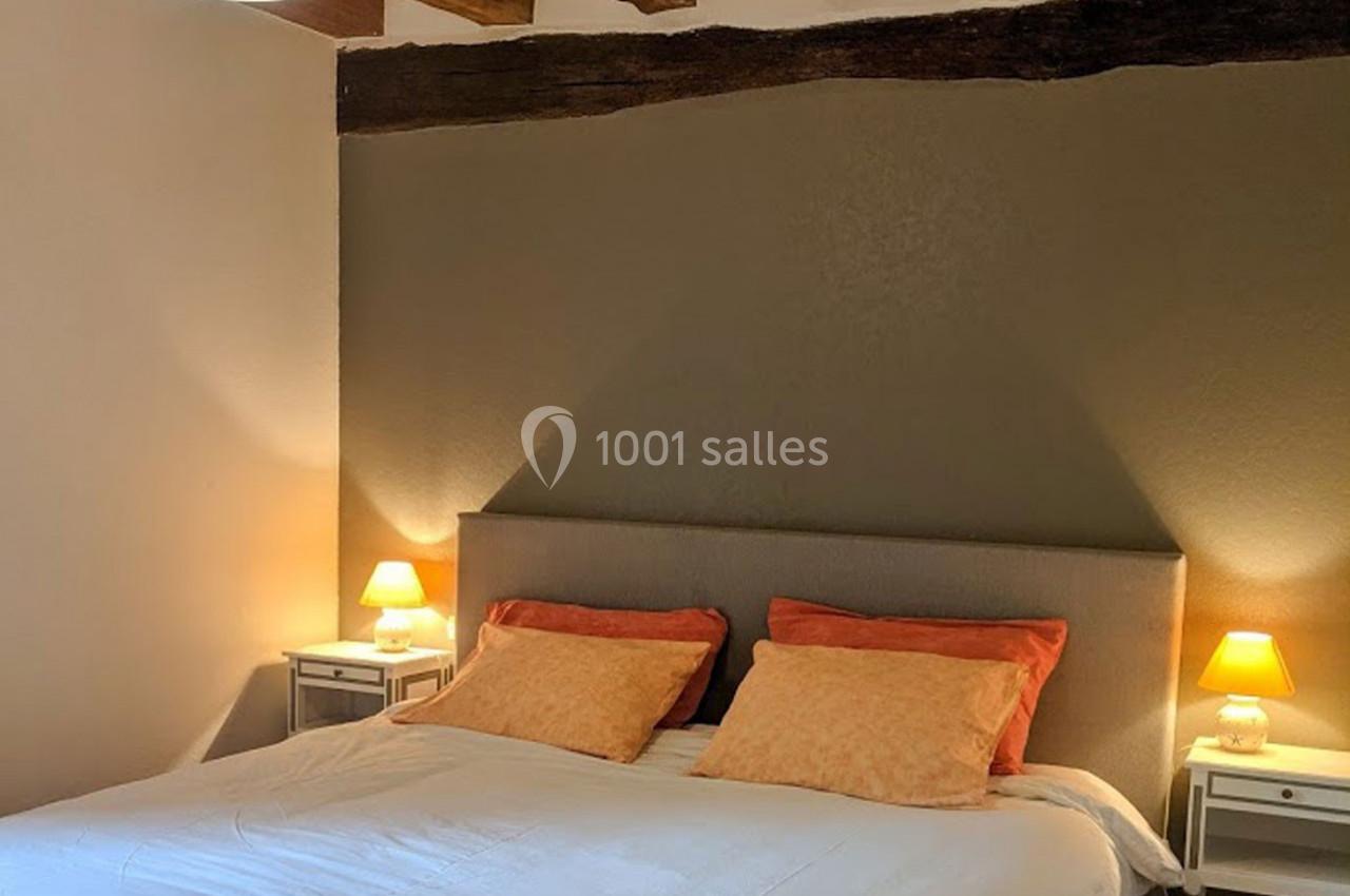 Lit double avec tête de lit grise, deux tables de chevet blanches et lampes allumées dans une chambre aux poutres apparentes.