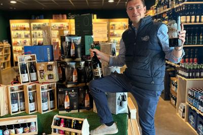 Un homme pose dans un magasin de boissons, tenant deux bouteilles devant un présentoir de bières variées.