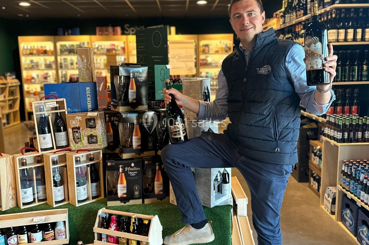 Un homme pose dans un magasin de boissons, tenant deux bouteilles devant un présentoir de bières variées.