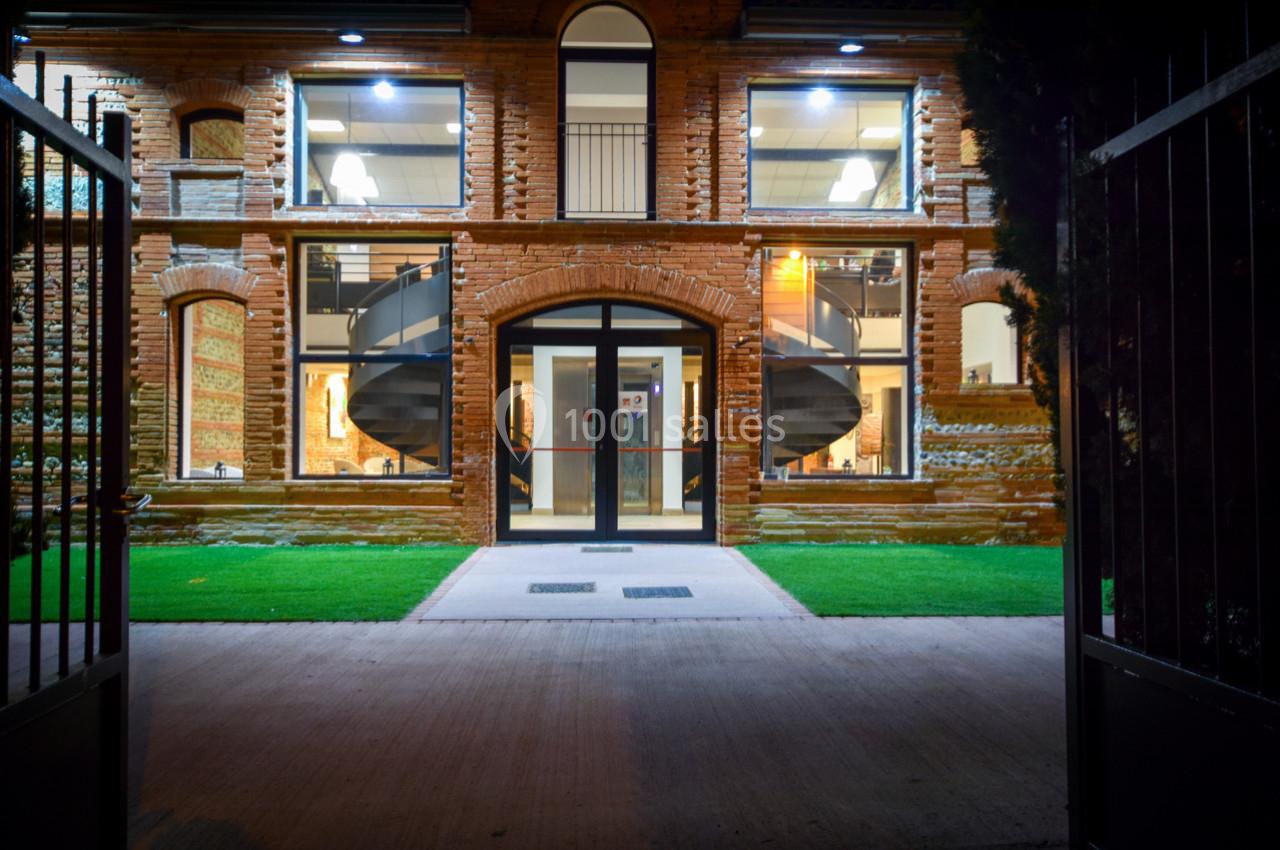 Façade d'un bâtiment en briques avec grandes fenêtres éclairées et escaliers en colimaçon visibles à l'intérieur.