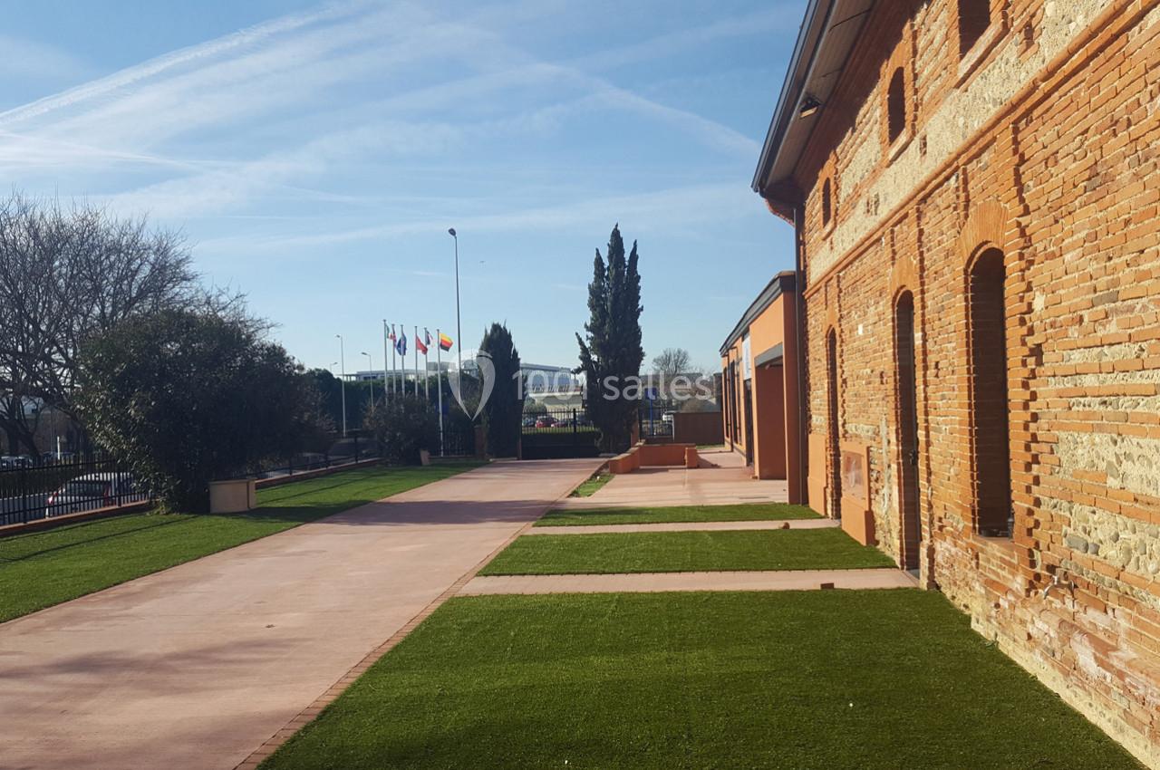 Bâtiment en briques rouges avec une allée bordée de pelouses et des drapeaux visibles au loin sous un ciel dégagé.