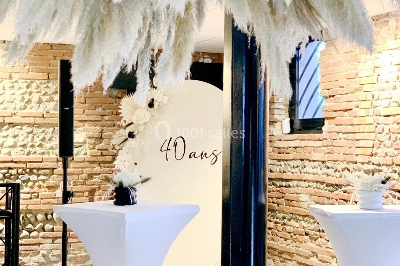 Décoration d'anniversaire avec des herbes de la pampa suspendues, murs en briques et tables hautes blanches.