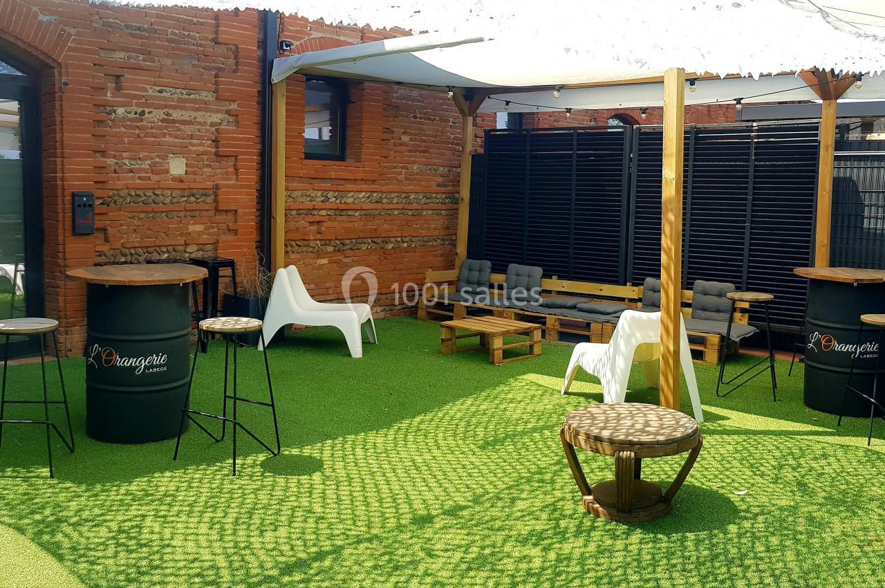 Espace extérieur aménagé avec tables hautes, chaises, canapés et parasol sur sol en gazon synthétique.