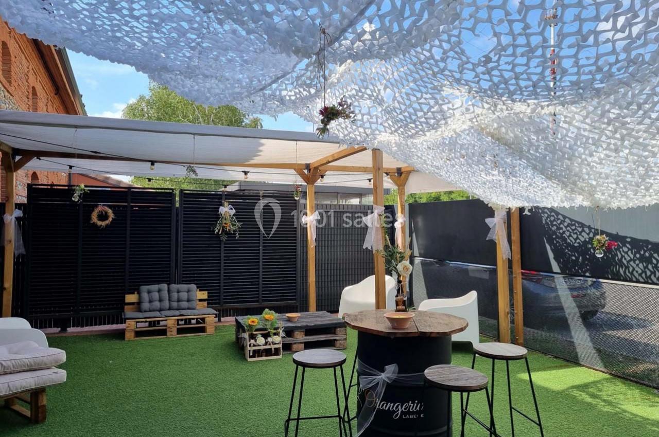 Espace extérieur aménagé avec des tables hautes, des fauteuils, un sol en gazon synthétique et une décoration suspendue.