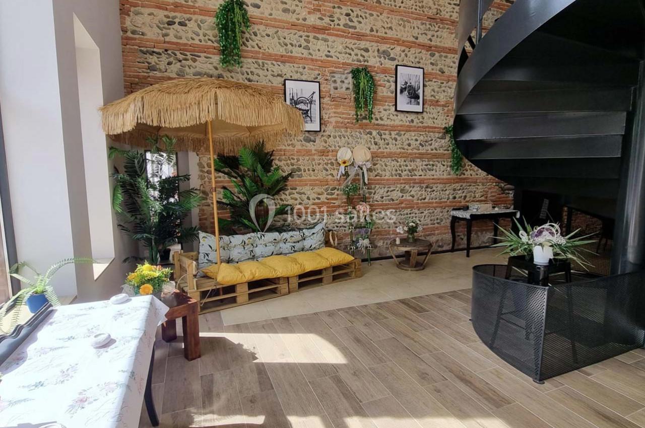 Espace intérieur lumineux avec canapé en palettes, parasol en paille, plantes vertes et escalier en colimaçon noir.