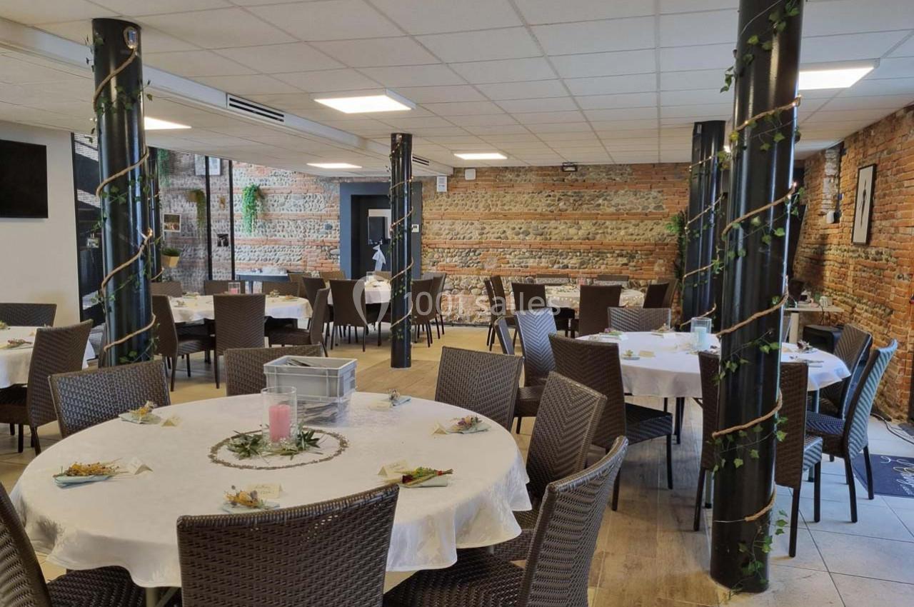 Salle de réception lumineuse avec murs en briques, tables rondes dressées et colonnes décorées de guirlandes végétales.