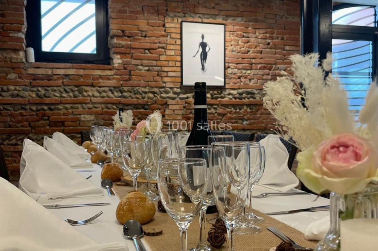 Table dressée avec nappes blanches, verres, pains et fleurs, dans une salle aux murs en briques apparentes.