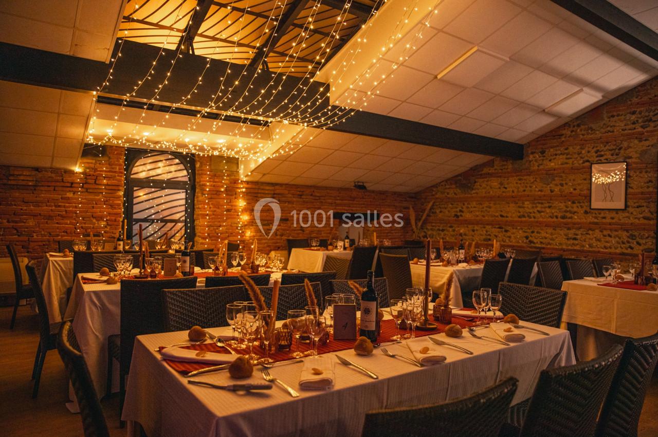 Salle de réception décorée avec des guirlandes lumineuses, tables dressées avec nappes blanches et vaisselle élégante.