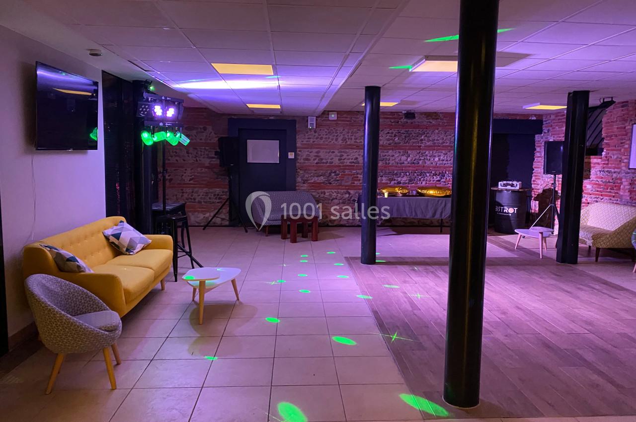 Salle avec un espace carrelé et parqueté, canapé jaune, tables basses, éclairages colorés et murs en briques.