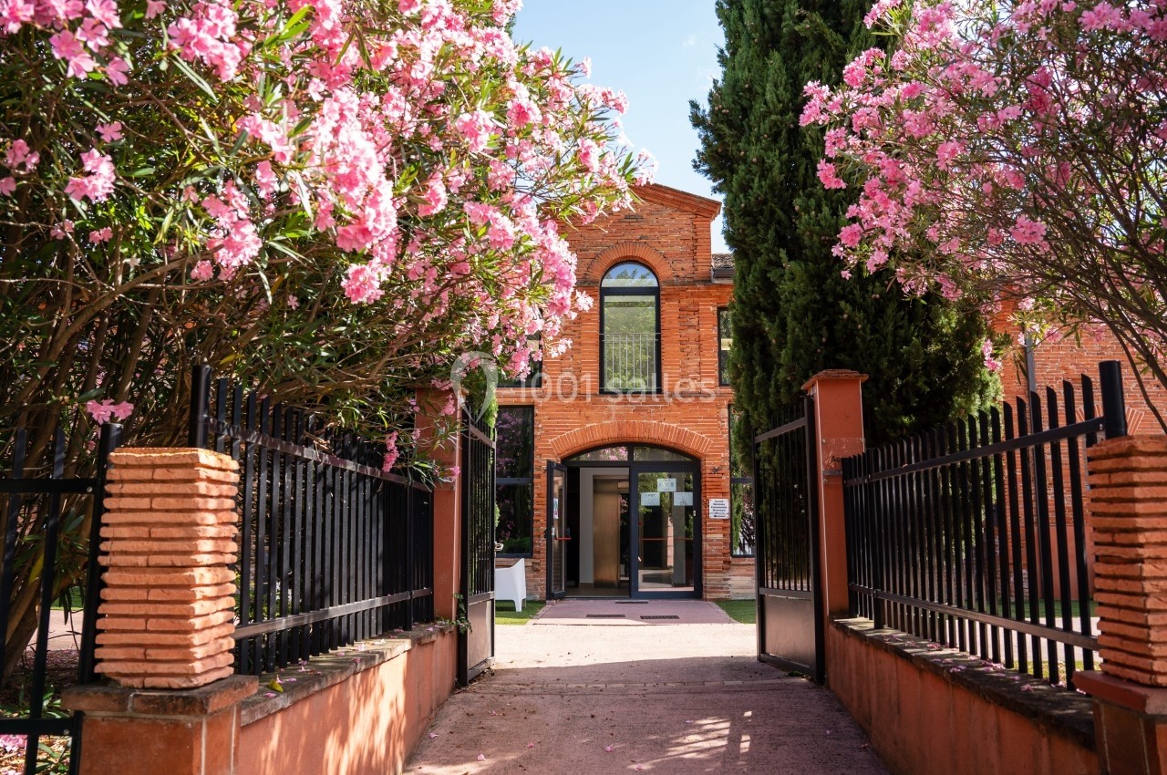 Entrée d'un bâtiment en briques rouges entourée de lauriers roses en fleurs et de grilles en fer forgé.