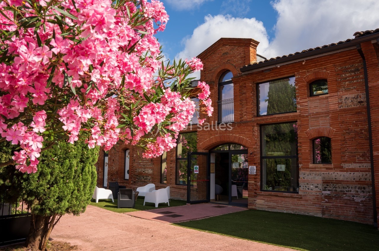 Façade en briques rouges d'un bâtiment avec des fenêtres cintrées, entouré de fleurs roses et de verdure.