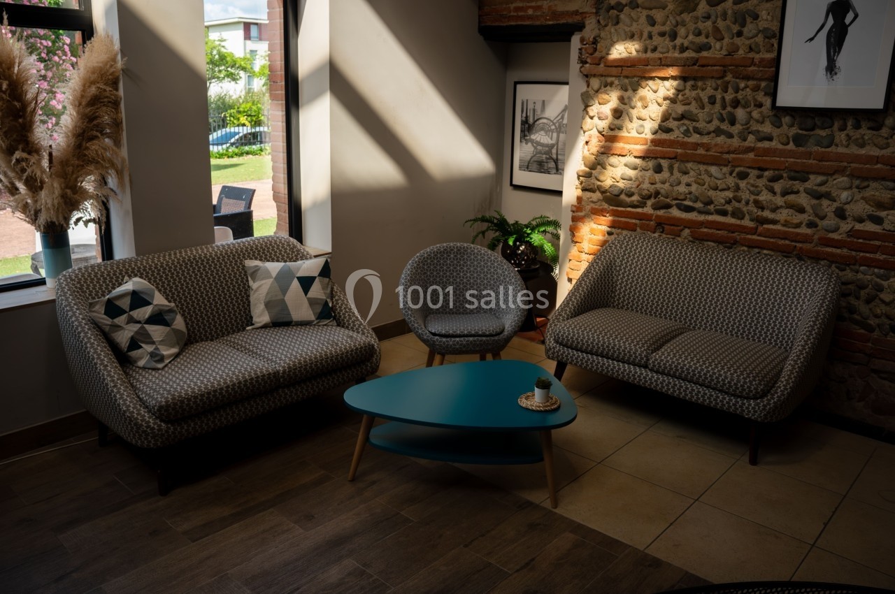 Salon lumineux avec canapé, fauteuil, table basse turquoise et mur en pierre, près d'une fenêtre.