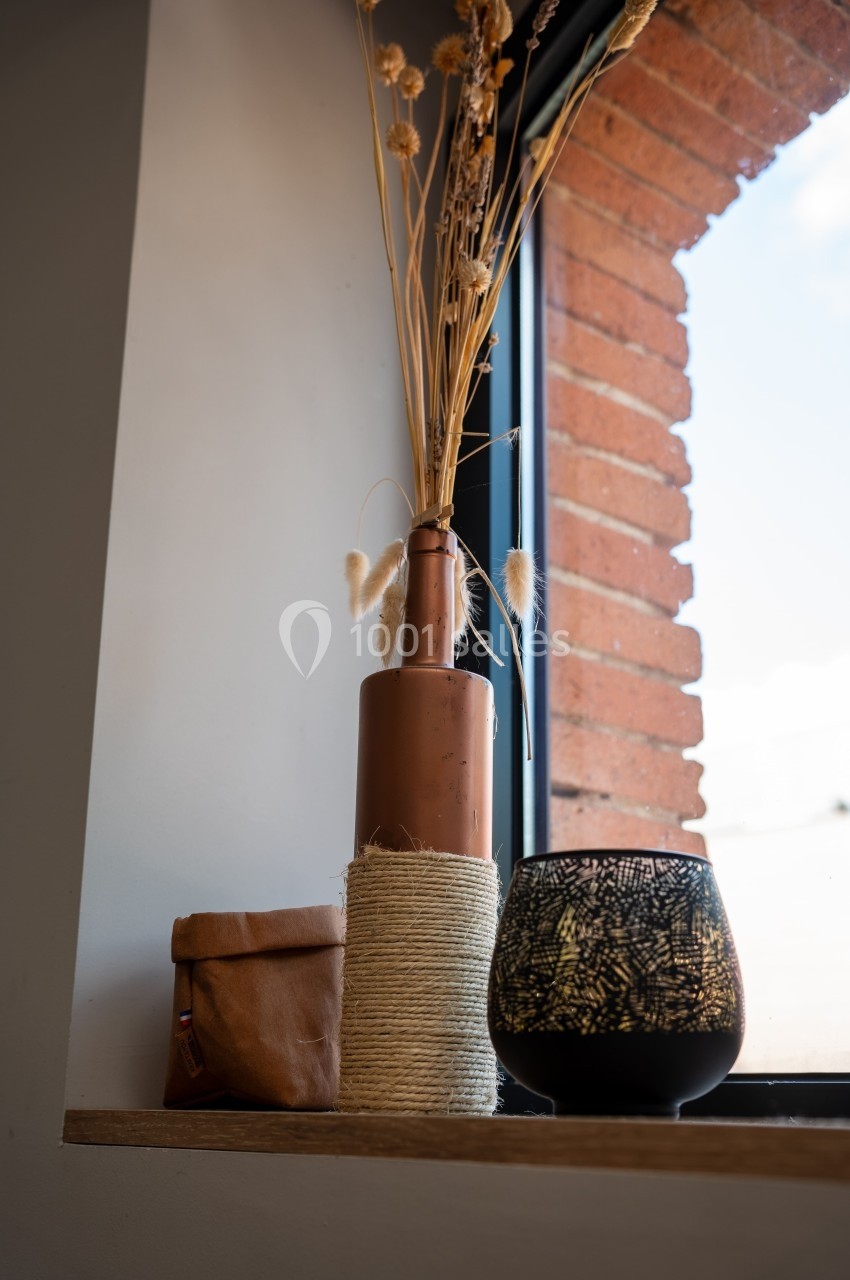 Vase en métal avec des herbes séchées, bougie décorative et pochette en tissu posés sur un rebord de fenêtre.