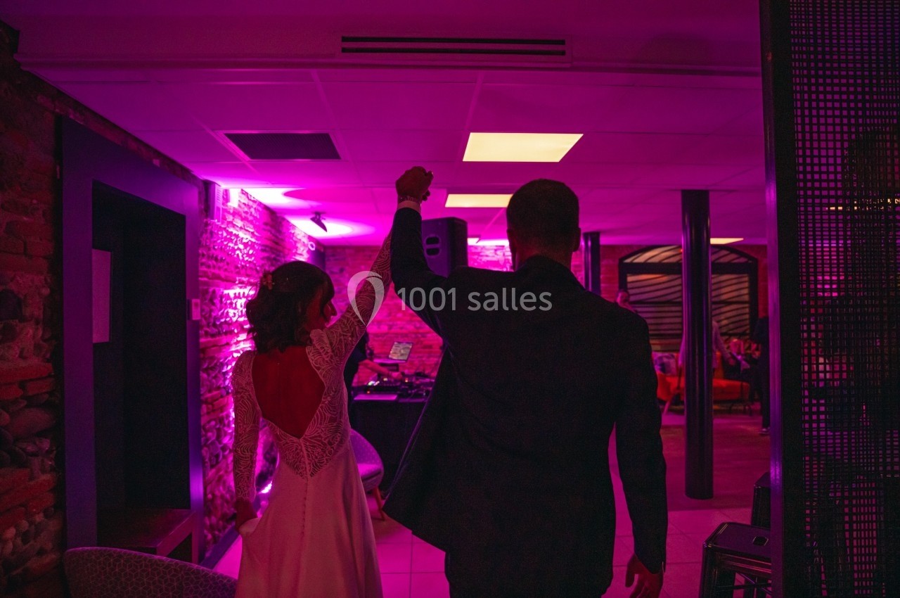 Un couple en tenue de mariage entre dans une salle éclairée par des lumières roses et violettes.