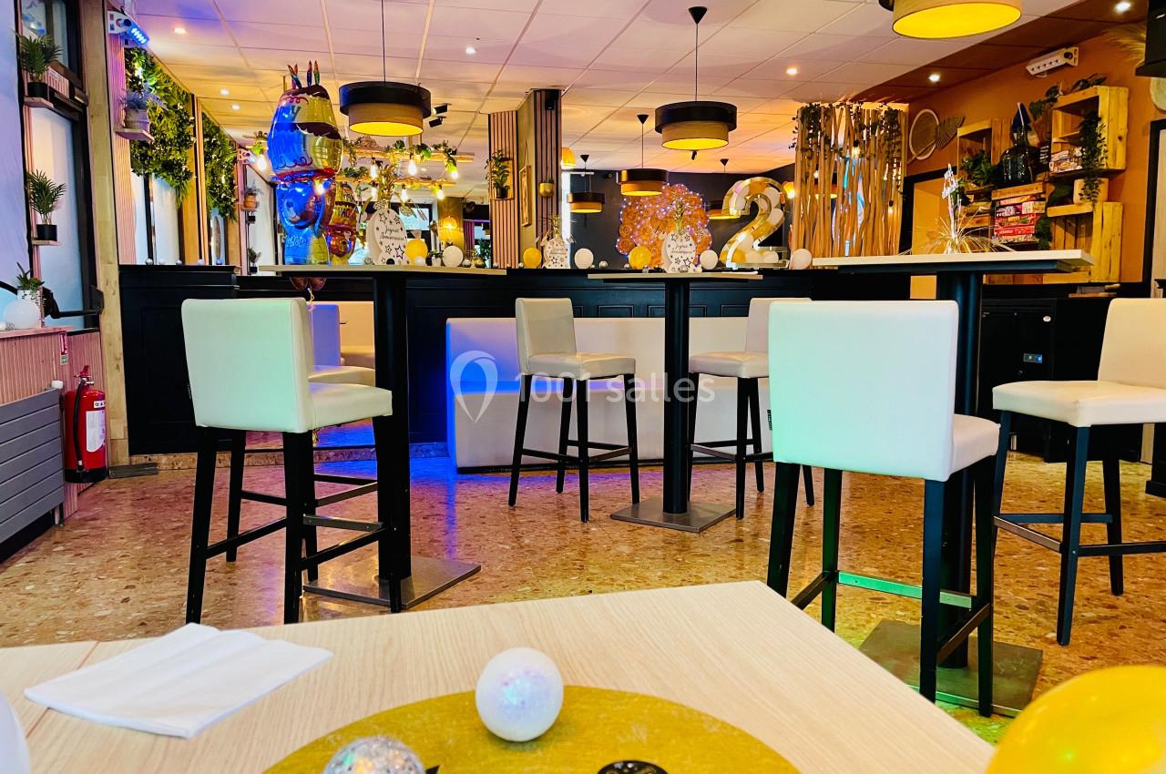 Salle décorée avec des tables hautes, chaises blanches et éléments festifs, éclairée par des suspensions lumineuses.