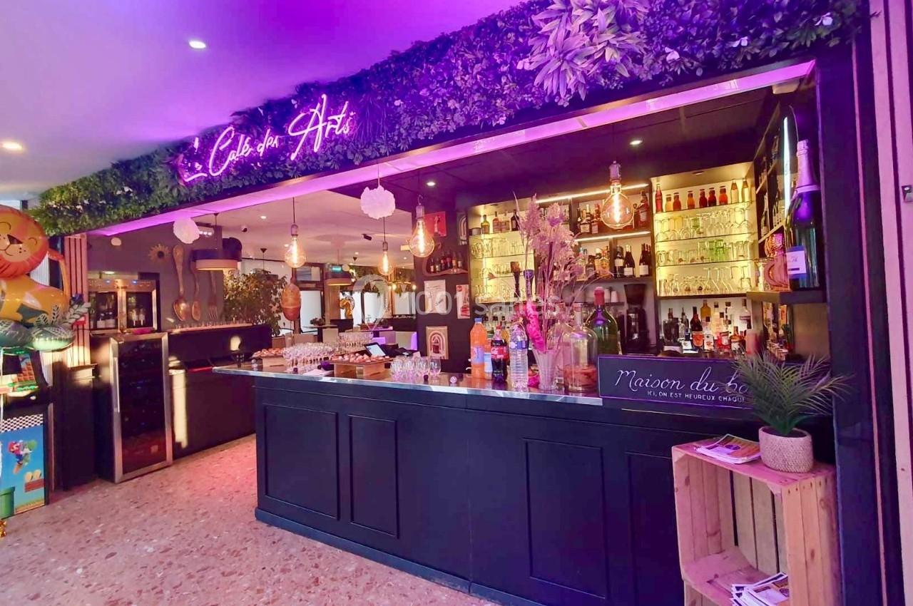 Bar décoré avec des plantes suspendues, éclairage néon violet, étagères de bouteilles et ambiance festive.