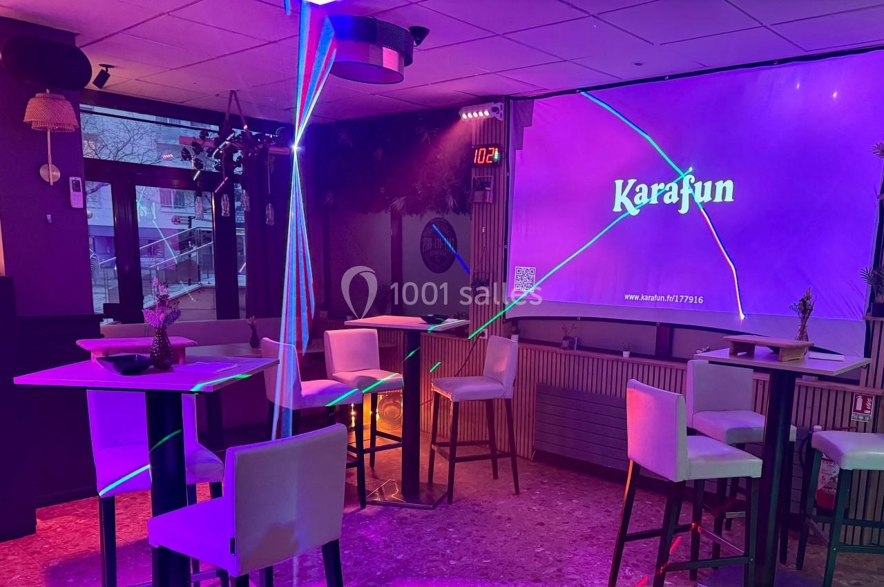 Salle de karaoké avec des tables hautes, des chaises blanches et un écran affichant ’Karafun’ sous des lumières colorées.