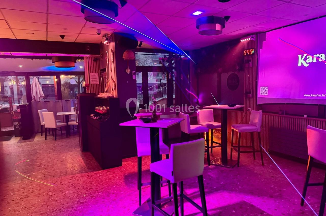 Salle de karaoké avec éclairage coloré, tables hautes, chaises blanches et vue sur une terrasse extérieure.