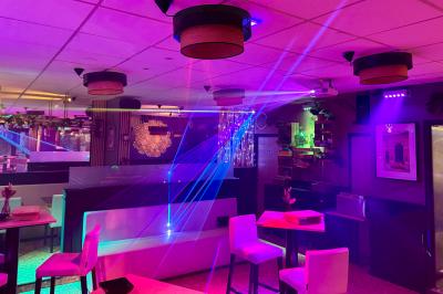 Salle de bar avec tables hautes, chaises, éclairage coloré et jeux de lumière dans une ambiance nocturne.