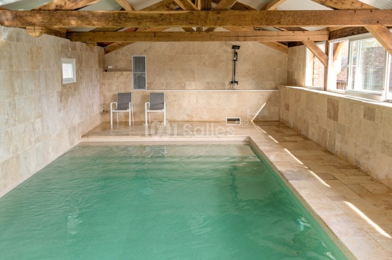 Piscine intérieure rectangulaire à l'eau turquoise, bordée de pierre beige, sous une charpente en bois apparente, avec deux chaises longues blanches en arrière-plan.