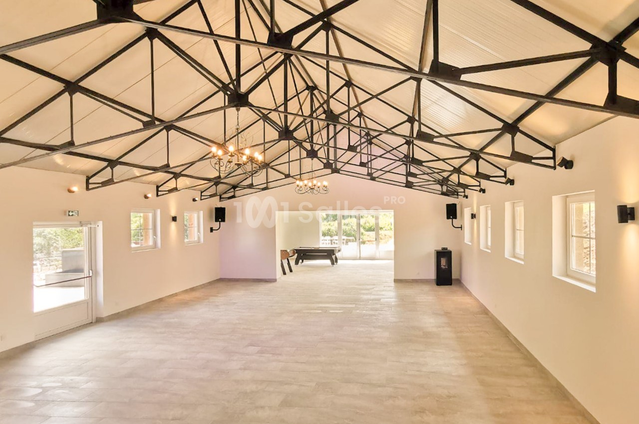 Grande salle vide au sol carrelage clair, charpente métallique apparente en noir, lustres en fer forgé et enceintes murales.