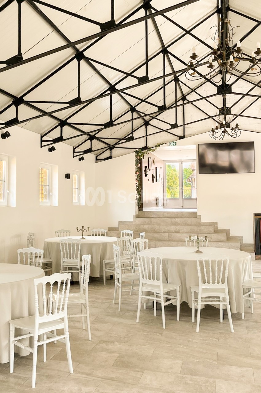 Grande salle lumineuse aux murs blancs, avec charpente métallique noire apparente, tables rondes nappées de blanc et chaises blanches dressées pour un banquet. Let me check length: roughly 165 characters, too long. Need ≤125. Salle lumineuse à charpente