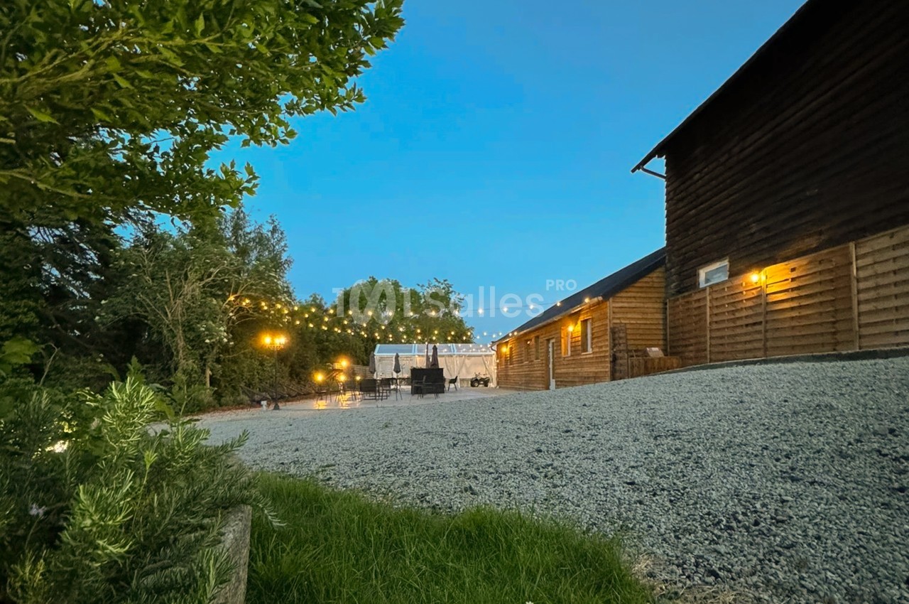 Cour en gravier au crépuscule bordée de bâtiments en bois éclairés par des guirlandes lumineuses, avec des tables et chaises sous un grand chapiteau blanc au fond.