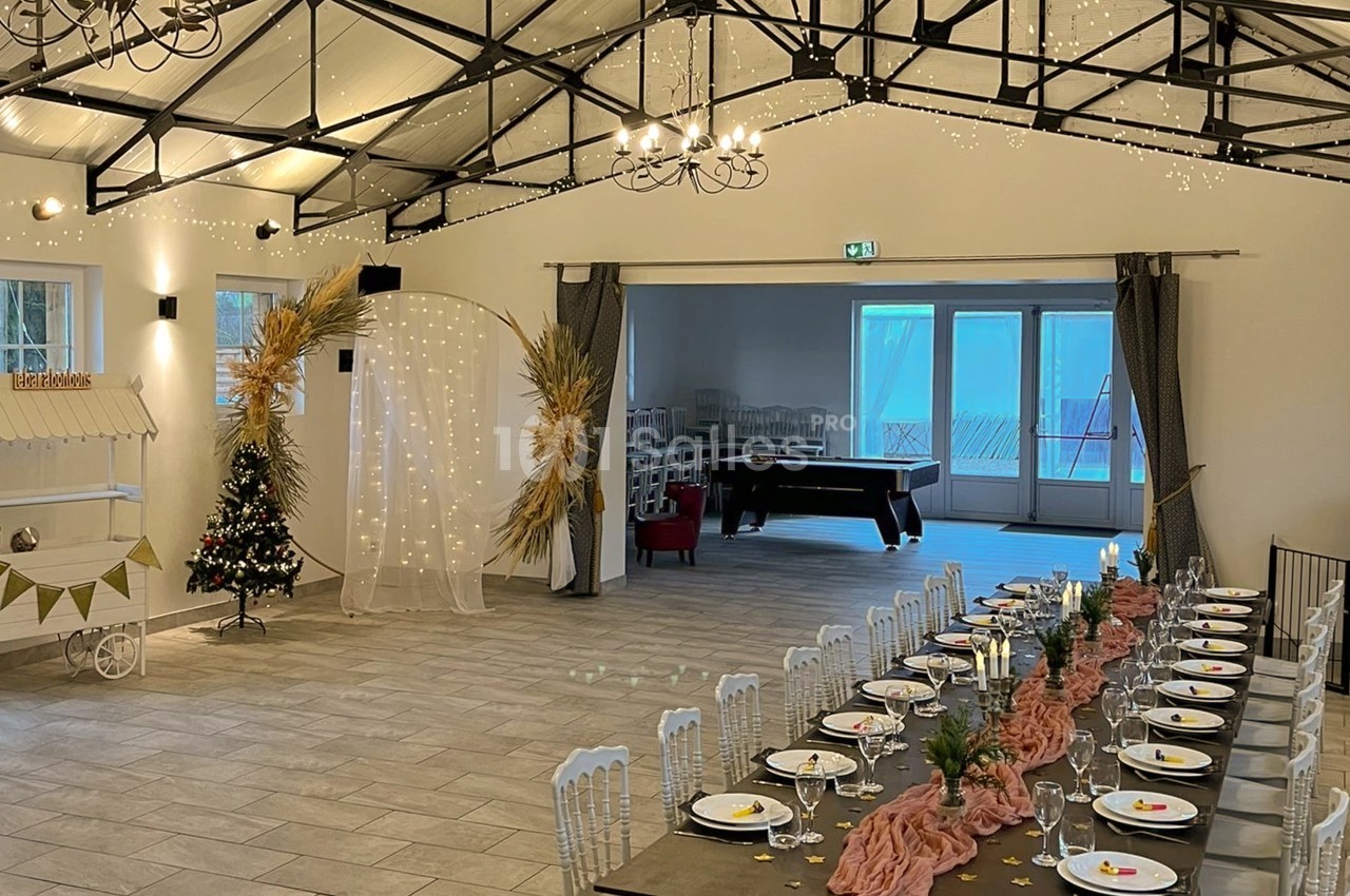 Grande salle lumineuse sous charpente métallique ornée de guirlandes, avec une table de réception dressée de nappes roses et de couverts blancs, et une billard visible au fond. Let me check constraints: under 125 chars? Count roughly: "Grande salle