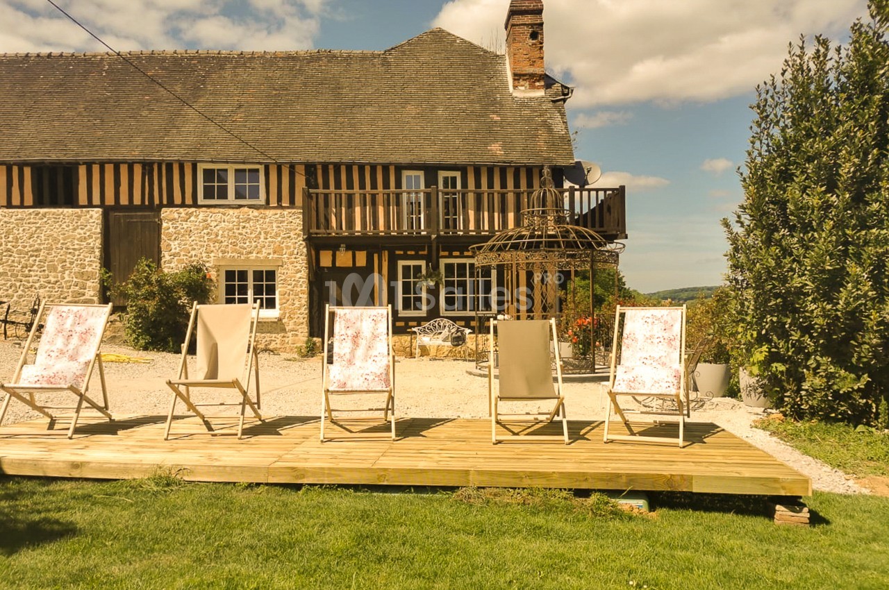 Maison normande à colombages avec terrasse en bois ensoleillée, munie de quatre transats au tissu fleuri et de deux chaises beiges. Let me check: count characters. "Maison normande à colombages avec terrasse en bois ensoleillée, munie de quatre transats