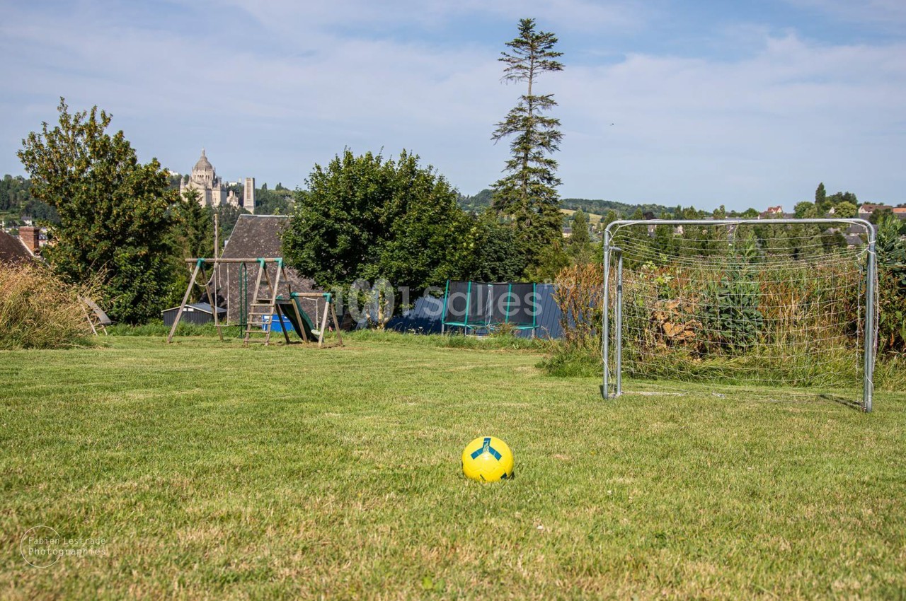 Pelouse de jardin avec un ballon jaune, un but de football, une balançoire et un trampoline sous un ciel clair.