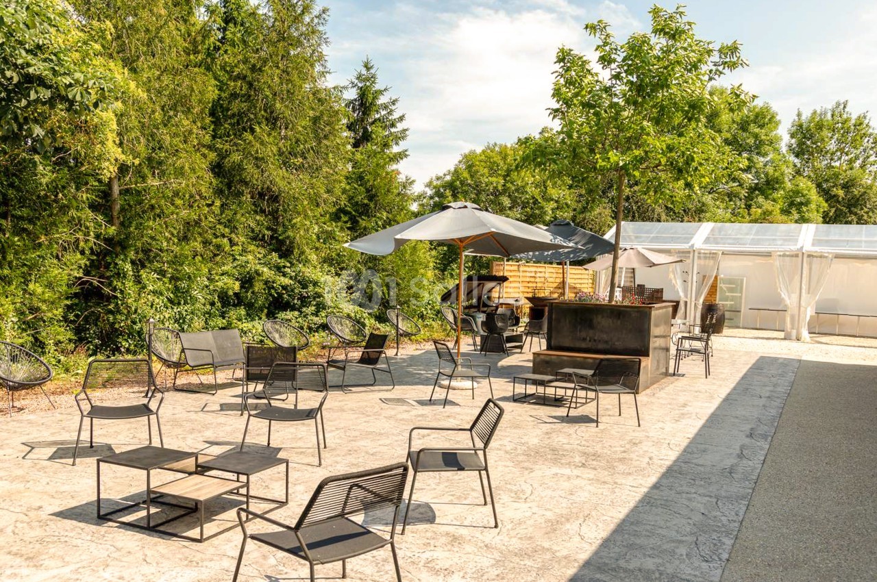 Terrasse en béton imprimé aménagée de fauteuils et tables basses en métal noir, sous des parasols, avec un bar en bois et métal et une structure blanche translucide à droite.